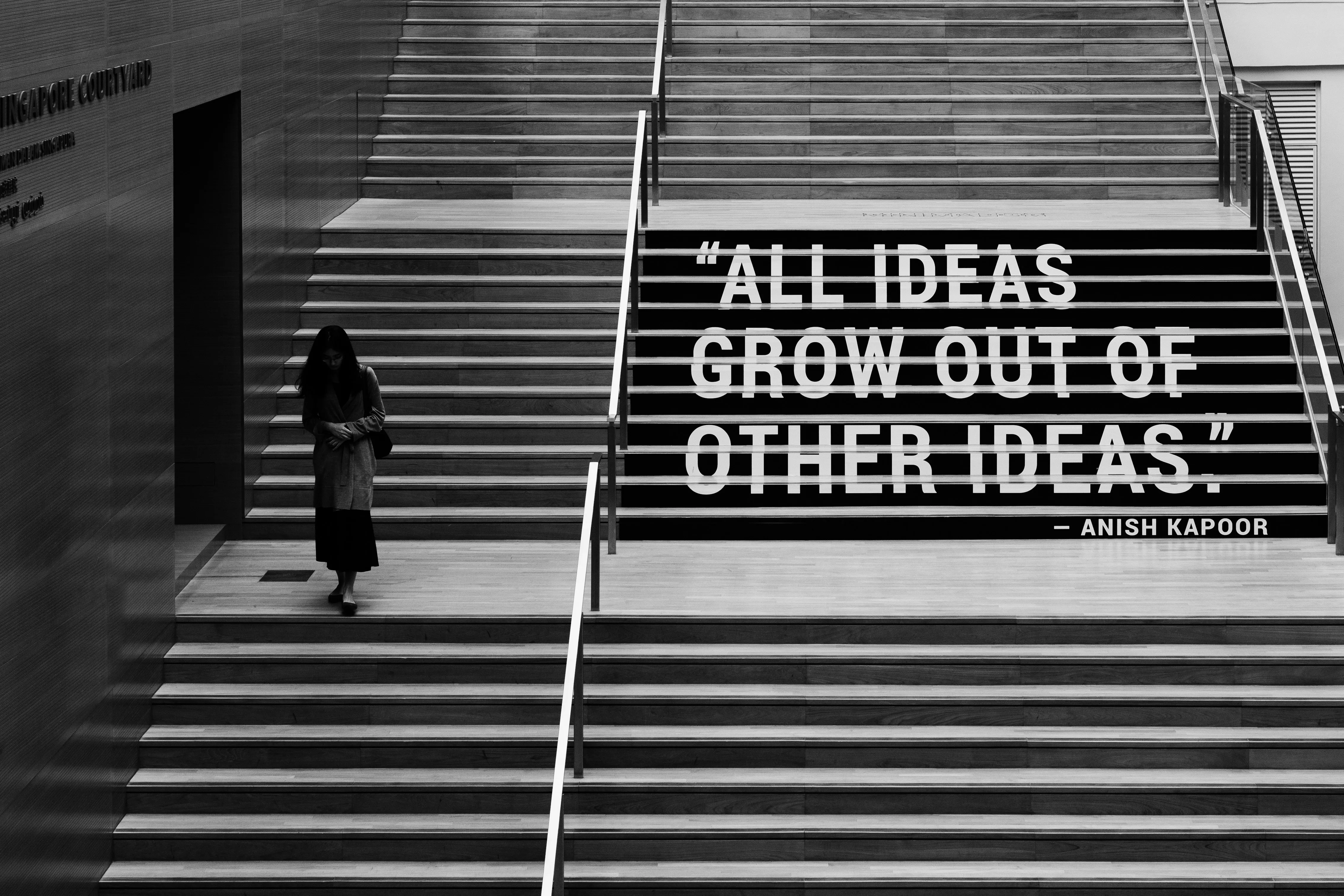 Person geht eine breite Treppe hinab, auf deren Stufen der Schriftzug All ideas grow out of other ideas von Anish Kapoor steht, Schwarzweiß-Aufnahme